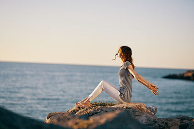 Young woman on rock by sea against clear sky