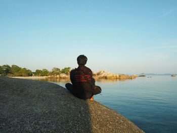 Rear view of woman sitting on rock by sea against clear sky