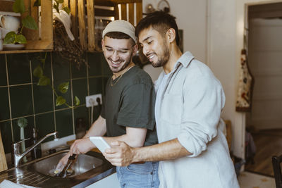 Smiling man sharing smart phone with boyfriend doing dishes in kitchen at home