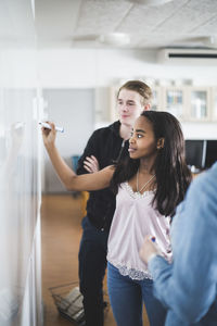 Confident girl writing on whiteboard while standing amidst teacher and friend in classroom