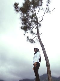 Side view of young man standing by tree against sky