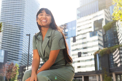 From below happy young asian female with long hair smiling and looking at camera while sitting against modern skyscrapers on sunny day on city street