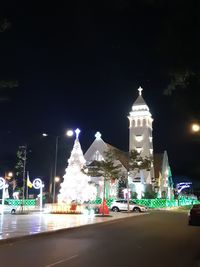 Illuminated street by buildings against sky at night