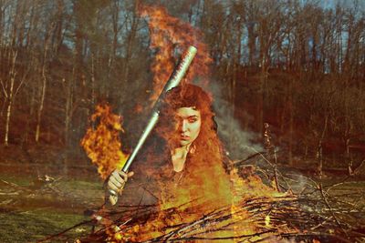 Double exposure of woman carrying baseball bat and fire burning in forest