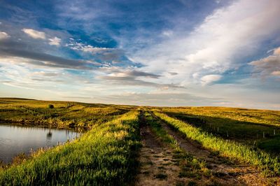 Dirt road on field against cloudy sky