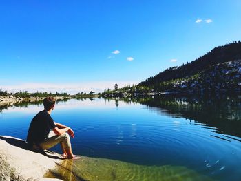 Man sitting on lakeshore against blue sky