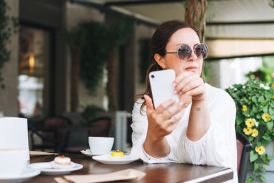 Beautiful middle aged woman in sunglasses in white clothes using mobile phone with coffee at cafe