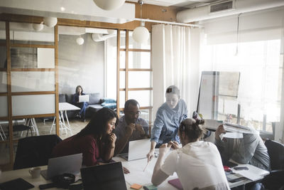 Male and female coworkers discussing businessman in meeting at office seen through glass wall