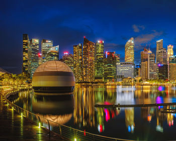 Illuminated modern buildings by river against sky at night