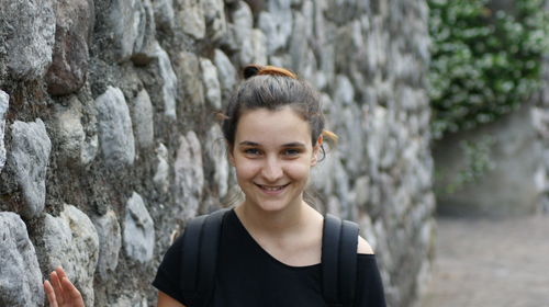 Portrait of young woman smiling while standing against stone wall