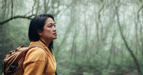 Side view of female hiker with backpack looking away while standing in forest during foggy weather