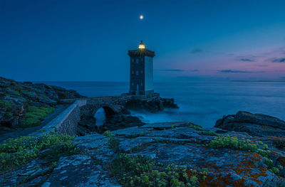 Lighthouse amidst sea and buildings against sky at dusk