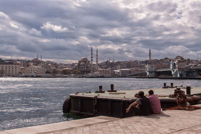 Rear view of friends sitting at harbor by mosques in city against cloudy sky
