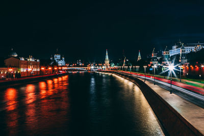 Light trails on river by illuminated city against clear sky at night