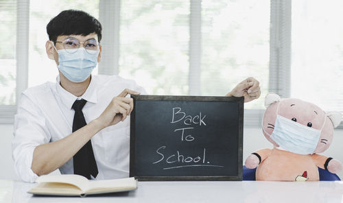 Portrait of man wearing flu mask holding slate sitting at classroom