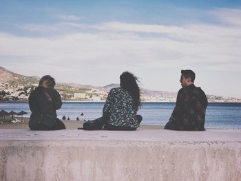 Friends sitting on retaining wall by sea against sky