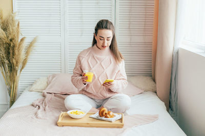 Smiling woman using mobile phone while sitting on bed by breakfast at home