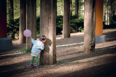 Full length of girl with balloon hiding behind tree trunk while playing at park