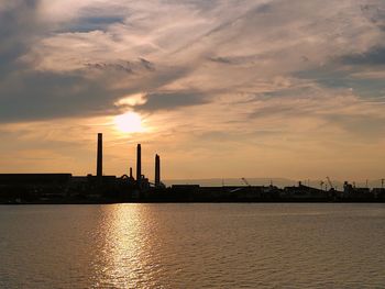 Silhouette factory by sea against sky during sunset