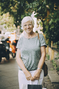 Portrait of smiling senior woman with bag standing on sidewalk in city