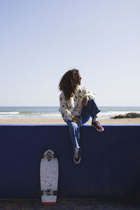 Full body of positive female sitting on handrail near skateboard against sandy waterfront with sea in sunny weather