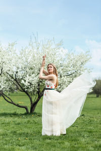 A young blonde in a long white dress poses near a cherry blossom in the garden, a spring landscape.