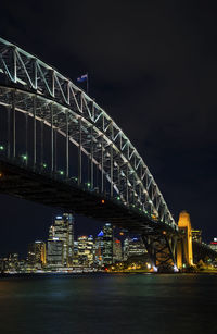 Illuminated bridge over river by buildings against sky at night