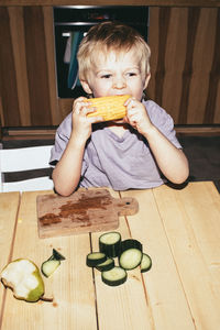 Hungry boy biting corn while sitting at table in home