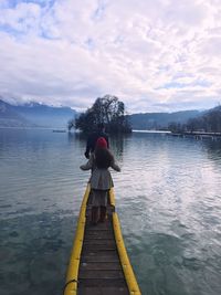 Rear view of man and woman standing on jetty over lake against cloudy sky