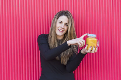 Portrait of smiling young woman holding drink in mason jar against wall
