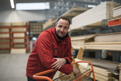 Portrait of confident salesman leaning on trolley in warehouse at hardware store