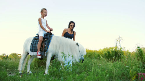 A woman and a boy are walking around the field, son is riding a pony, mother is holding a pony for