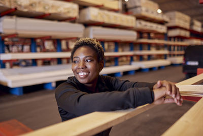 Smiling female worker looking away while leaning on plank in lumber industry