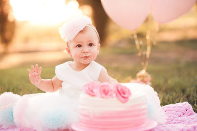Baby girl 1 year old eating creamy birthday cake sitting on green grass with pink balloons outdoors