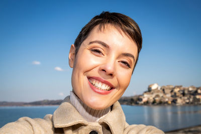 Beautiful young woman with short hair is taking a selfie in front of the lake.