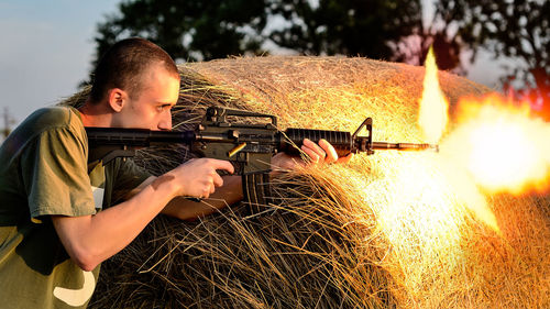 Side view of young man shooting with rifle against hay bale