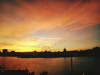 Silhouette buildings by river against sky during sunset