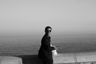 Portrait of smiling young woman standing on retaining wall against sea