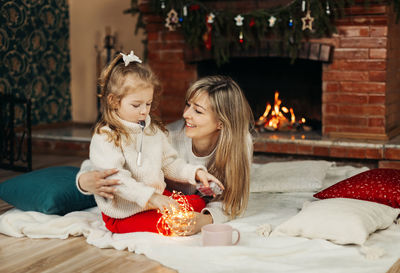 Mom and daughter are playing with christmas garlands on the floor by the fireplace