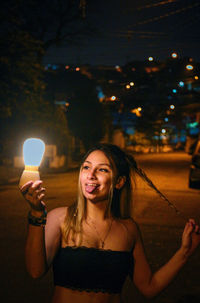 Portrait of smiling young woman holding illuminated lighting equipment at night