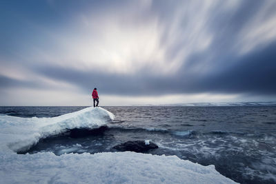 Man standing on snow covered rock by sea against cloudy sky