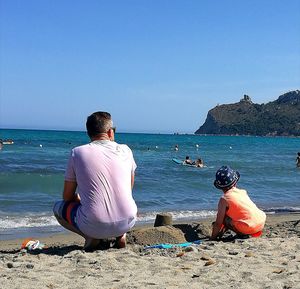 Rear view of man sitting on beach against clear sky