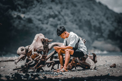 Full length of man sitting on driftwood at land