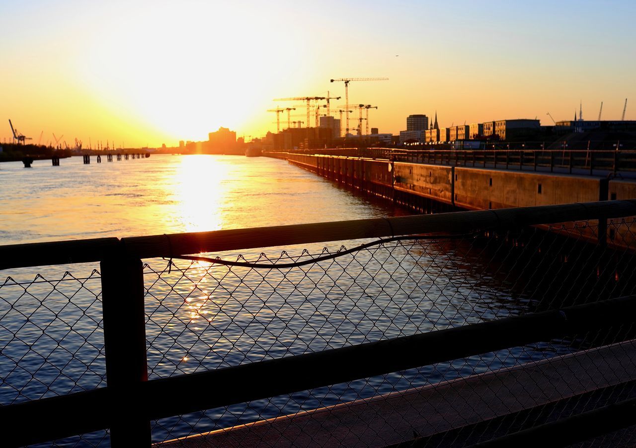 BRIDGE OVER RIVER AGAINST BUILDINGS DURING SUNSET