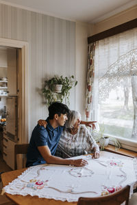 Male healthcare worker gesturing while sitting with senior woman near window at home