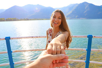 Cropped image of boyfriend holding girlfriend hand against lake and mountains during sunny day
