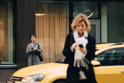 Businesswomen using smart phones while standing on street against building in city