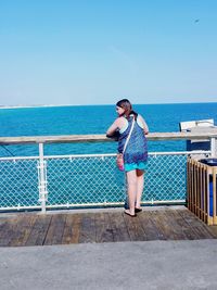 Rear view of woman leaning on railing at pier by sea against clear blue sky