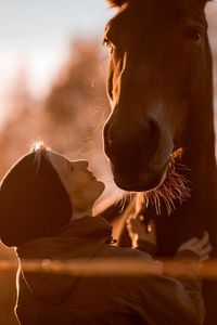 Side view of mid adult woman stroking horse while standing against sky in barn during sunset