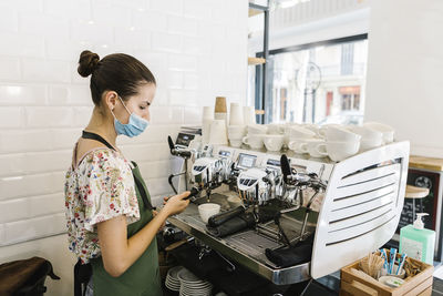 Young woman with face mask making coffee while standing in kitchen at coffee shop